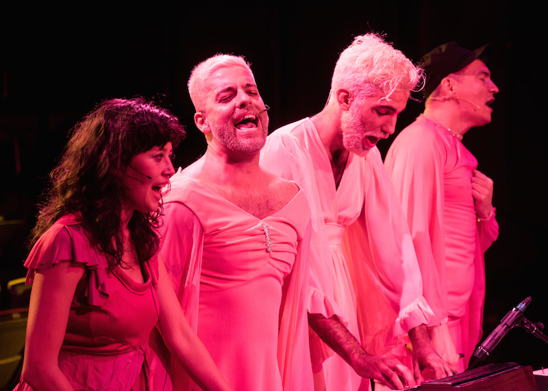 The performers of Part 2, cast in a pink light, sing towards the audience